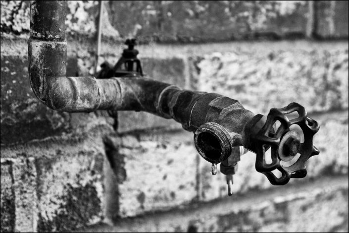 Close-up black-and-white image of a hose bibb on a textured brick wall, with a water droplet suspended at the spigot's tip, capturing a moment of stillness and anticipation.