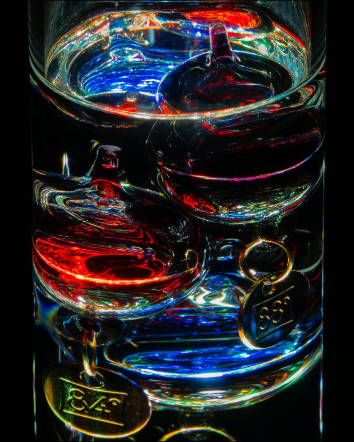Close-up photograph of a Galileo Thermometer with glowing orbs suspended in glass, captured with backlighting against a black background to highlight its colorful, jewel-like details and intricate design.