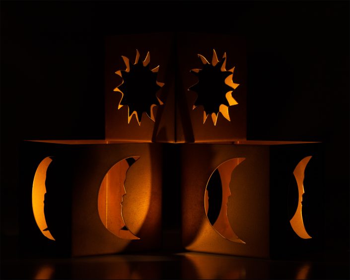 Photograph of three brass candle boxes with intricate cutouts, illuminated against a dark background, highlighting the interplay of light and shadow in a contemplative composition.