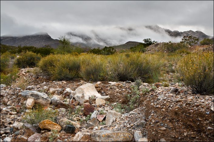 View of the Franklin Mountains' northeast foothills in El Paso, Texas, under a cloudy sky, featuring earthy desert tones, rocky textures, and a soft atmospheric haze that highlights the natural beauty and rugged landscape of the Southwest.