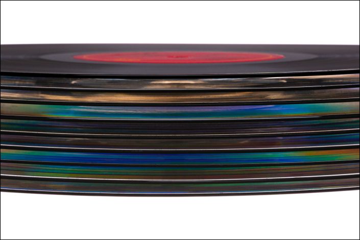 Features a Close-up arrangement of stacked vinyl records, highlighting their grooves and the interplay of light and color creating an abstract representation of sound and rhythm. Celebrates the beauty of vinyl while inviting reflection on the emotional depth of music.