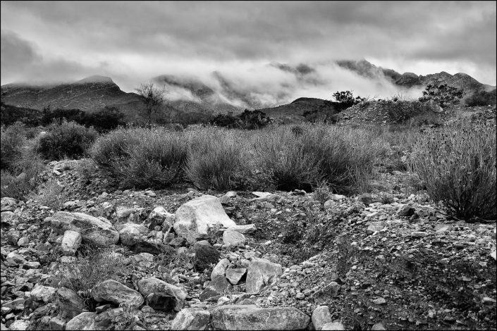 A black-and-white landscape of the Franklin Mountains in El Paso, Texas, features rugged foothills, cloud-covered peaks, and dramatic light and shadow contrasts.