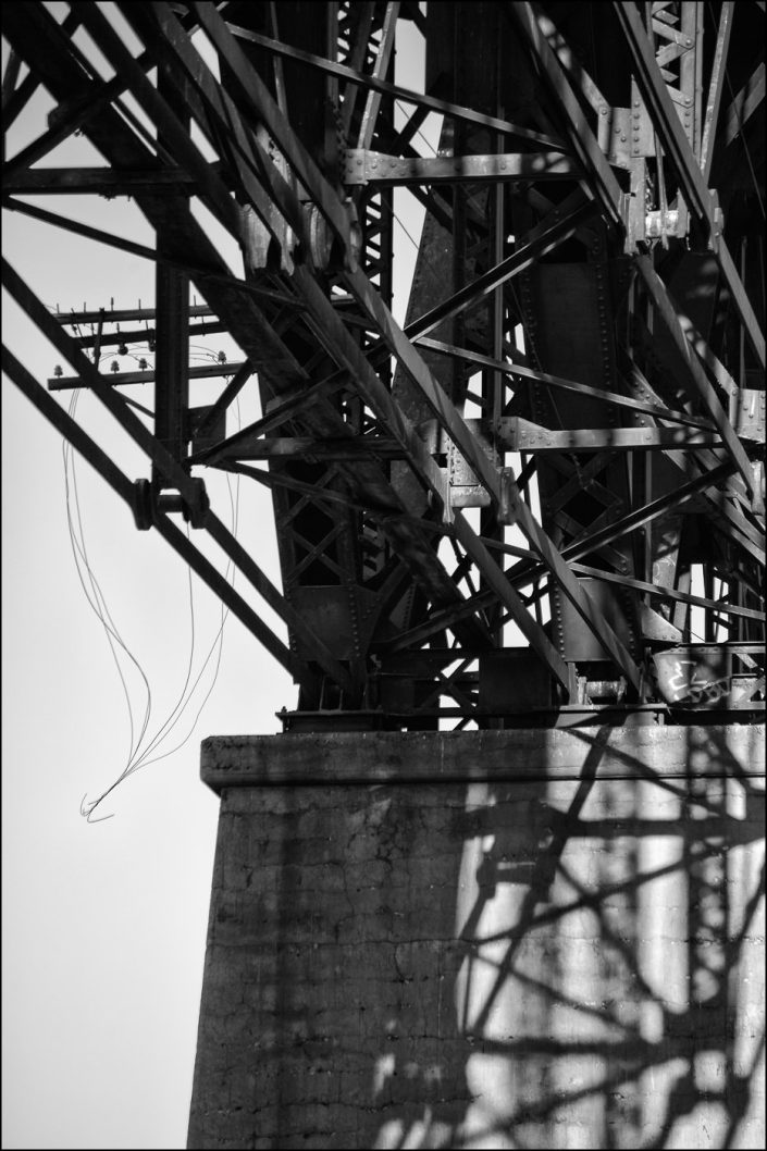 Black-and-white photograph of the underside of a Union Pacific bridge at the Texas–New Mexico border, featuring a complex network of steel girders and beams, with strong geometric patterns highlighted by light and shadow.