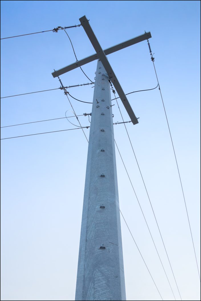 Minimalist photograph of a utility pole and power lines against a soft blue sky, emphasizing clean lines, contrast, and the abstract beauty of industrial design.