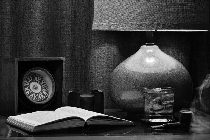 Black-and-white tableau featuring a vintage scene with a glass of scotch, smoldering pipe, open book, and antique compass, evoking solitude, reflection, and Hemingway-inspired ambiance in warm, ambient lighting.
